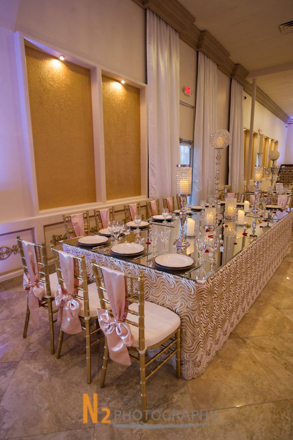 Elegant banquet table setup with mirrored tabletop, gold chairs adorned with pink satin bows, crystal candle holders, and fine china in a luxury event hall - Alegria Gardens - Houston, TX