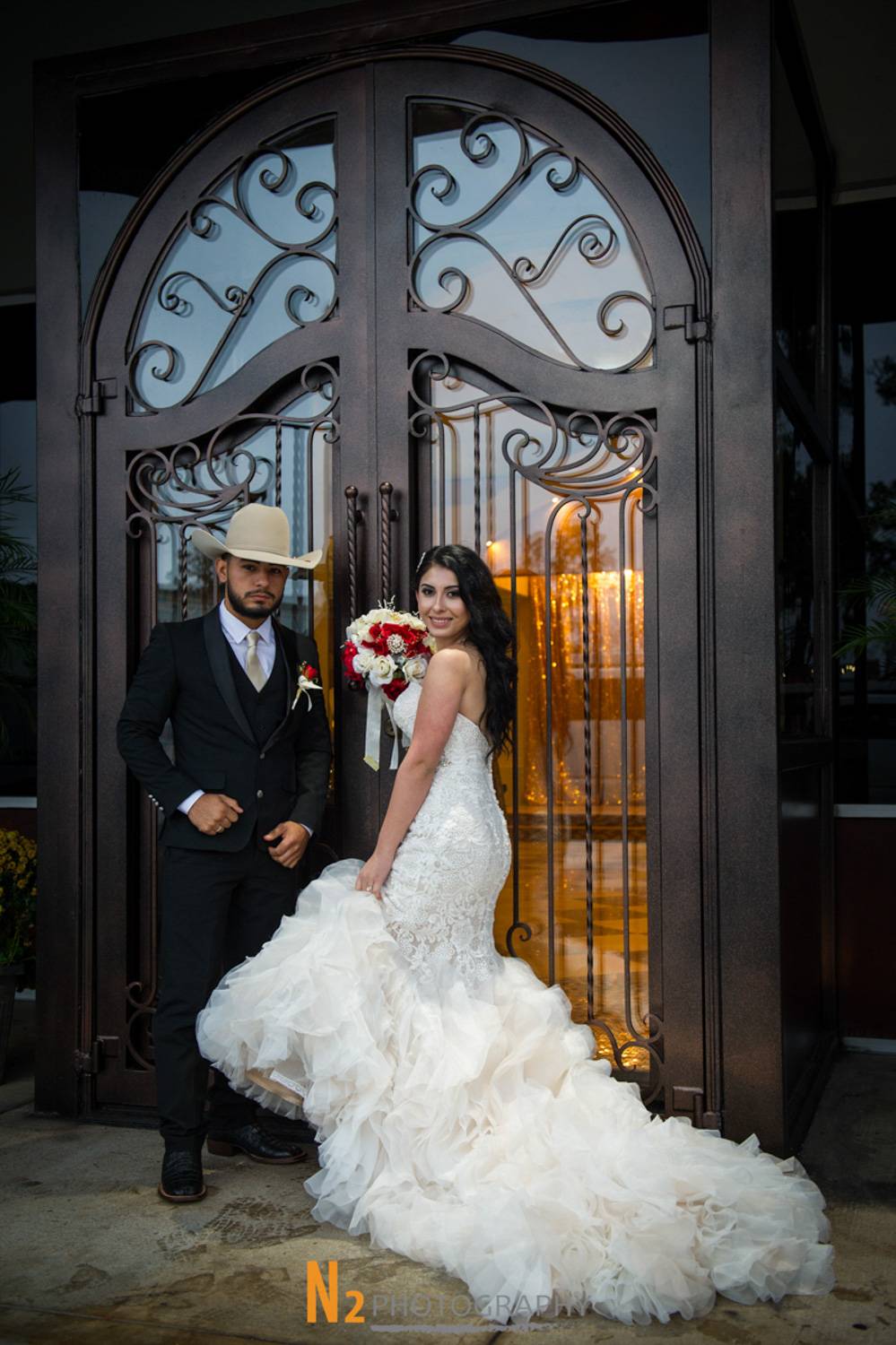 Bride and groom posing elegantly outside a venue with ornate wrought iron doors, showcasing timeless wedding style by Alegria Gardens event planners.
